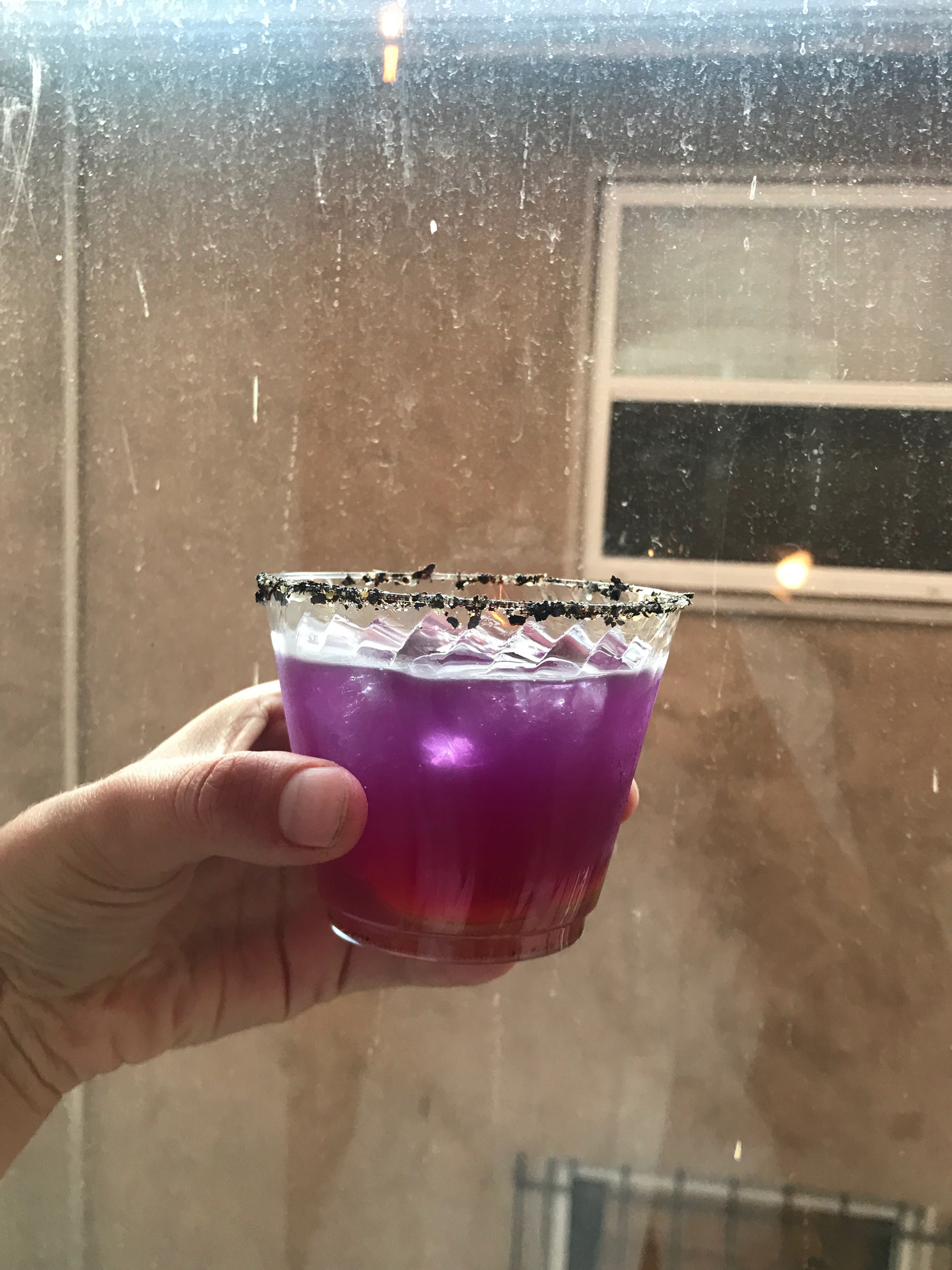 A hand holding a purple drink in a cleap cup in front of a window